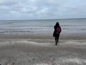 eine Frau spaziert im Wintermantel auf einem Sandstrand am Meer