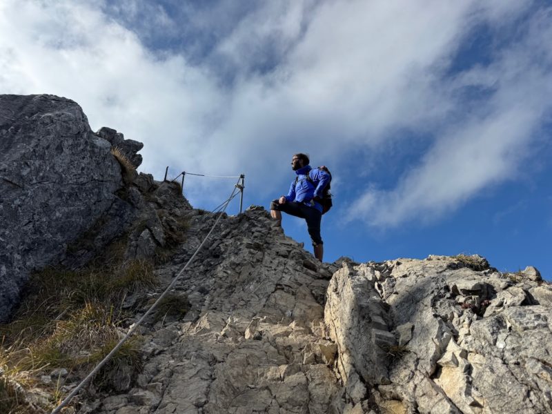 Wanderer steht auf felsigem Grat unterhalb des Gipfels der Brecherspitze, mit blauem Himmel und Sonne über den Alpen.