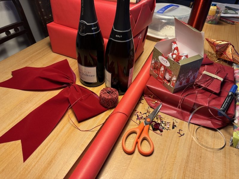 Geschenke werden an Heiligabend verpackt: rotes Geschenkpapier, Schleife, Schere, Garn und zwei Flaschen Champagner auf einem Tisch.