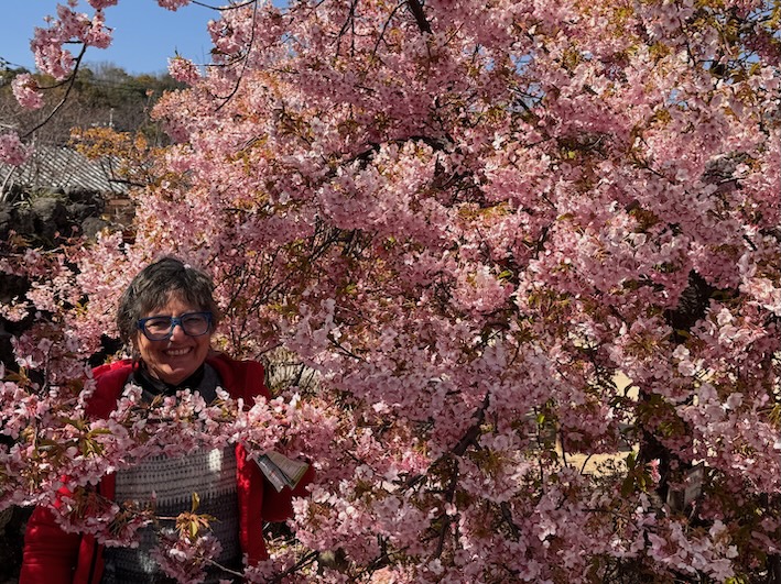 Frau steht lächelnd unter rosa Kirschblüten im Frühling – stimmungsvolles Bild zum Welttag der Poesie und einem persönlichen Gedicht über Erinnerungen und alte Worte.