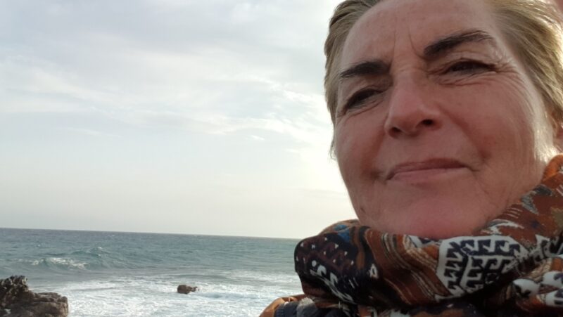 Nahaufnahme einer Frau am Meer bei windigem Wetter. Im Hintergrund sind Wellen und ein grauer Himmel zu sehen. Ihr Blick wirkt ruhig und nachdenklich.