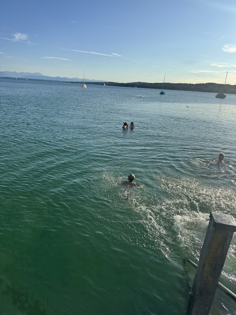 Menschen schwimmen in einem klaren See; im Hintergrund sind Boote und Berge unter blauem Himmel zu sehen.