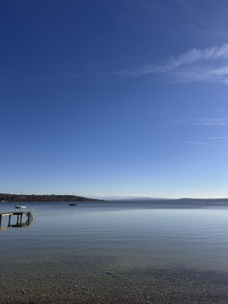 Ruhige Landschaft mit See, klarem Himmel und Steg am Ufer; ein Boot liegt auf dem Wasser, im Hintergrund sind entfernte Hügel zu sehen.