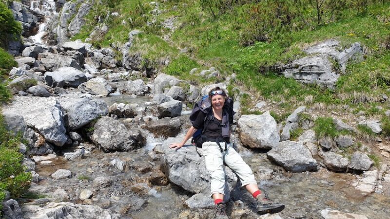 Frau sitzt lächelnd auf Felsen in einem Bergbach, umgeben von grüner Natur und Steinen. Sie wirkt entspannt und zufrieden auf ihrer Reise.
