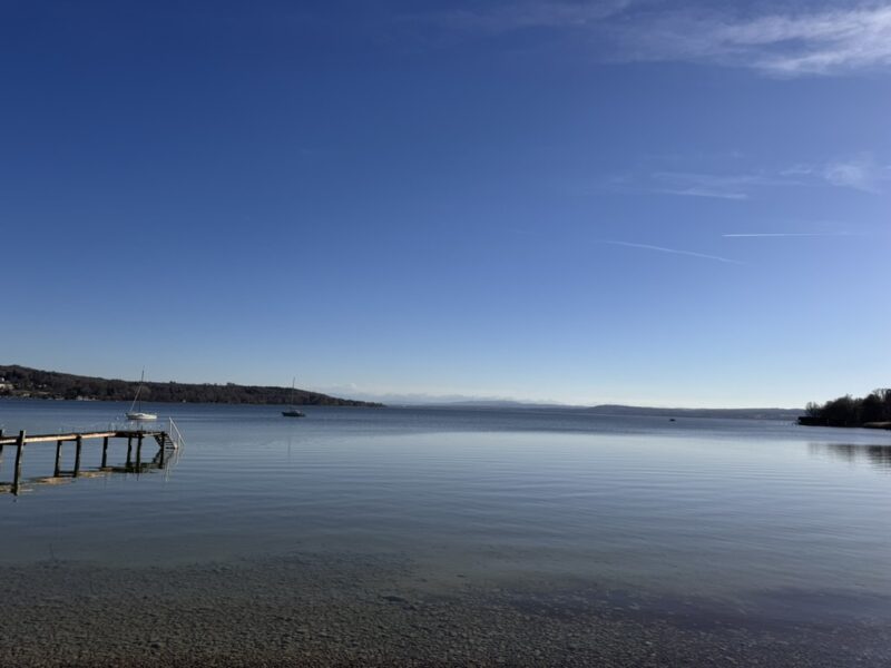 Ruhiger See mit klarem Wasser und weitem Himmel. Ein kleiner Steg ragt ins Wasser, im Hintergrund liegen Boote und eine sanfte Uferlandschaft.