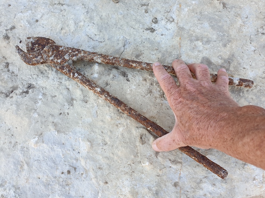 Hand hält eine alte, rostige Zange auf hellem Boden. Das Werkzeug wirkt abgenutzt und grob, die Szene ist nah aufgenommen.