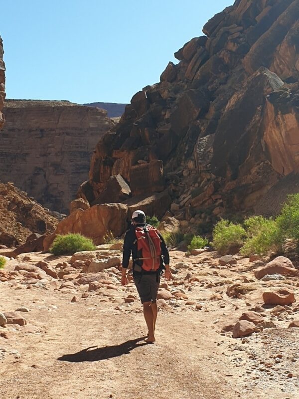 Wanderer mit Rucksack auf einem felsigen Weg durch einen roten Canyon. Das Bild symbolisiert Umwege, Orientierung und die Erkenntnis, dass Verirren manchmal zu den schönsten Erfahrungen einer Reise gehört.
