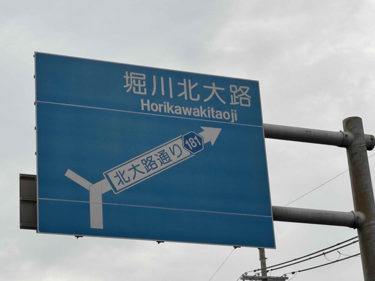 Blaues Straßenschild mit japanischer und lateinischer Beschriftung in Japan. Das Foto symbolisiert Orientierung, Fremde und den Beginn eines neuen Reiseabenteuers.