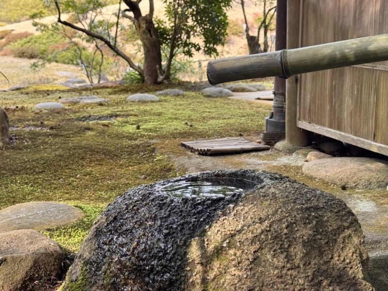 Bambusrohr über einem Stein im japanischen Zengarten, aus dem nur alle zehn Sekunden ein Wassertropfen fällt – Symbol für das japanische Konzept „Ma“, den wertvollen Zwischenraum und die Kraft der bewussten Pause.