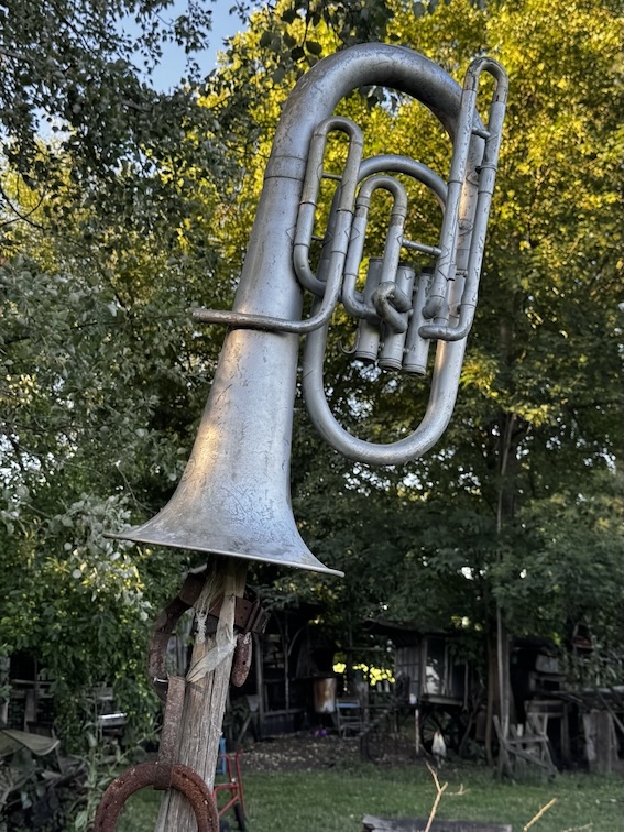 Großes silbernes Blasinstrument als Gartenskulptur zwischen grünen Bäumen