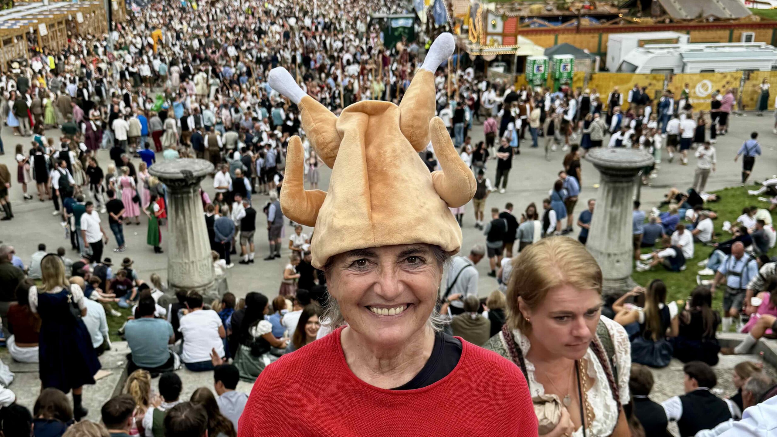 Autorin Irmgard Rosina Bauer mit lustigem Hendl-Hut vor einer großen Menschenmenge auf dem Oktoberfest in München.