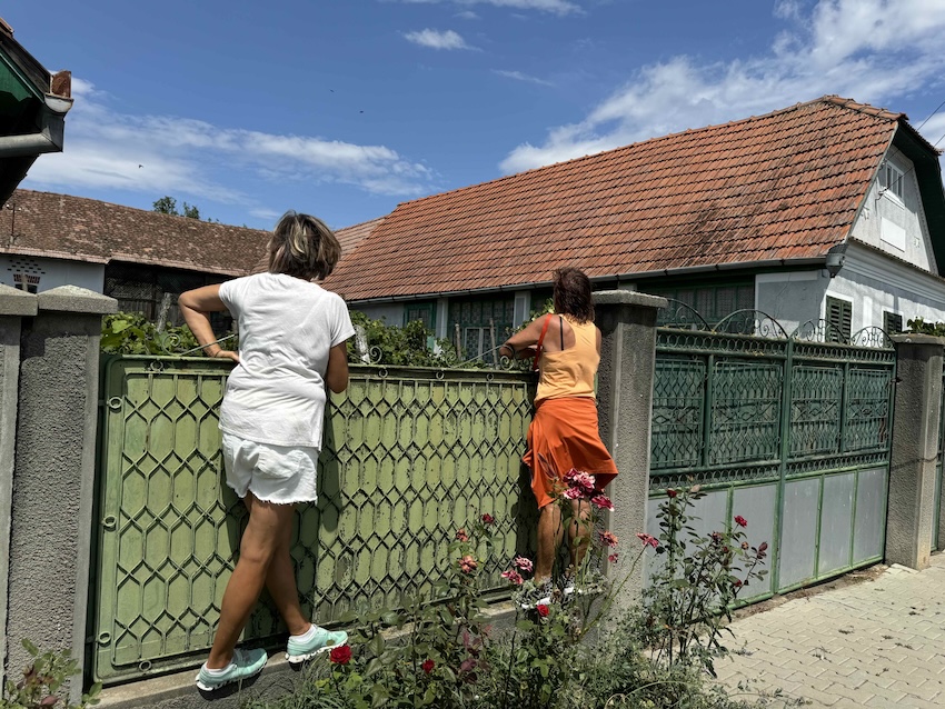 Zwei Frauen blicken über ein grünes Hoftor auf ein siebenbürgisches Haus – Sinnbild für Herkunft, Heimat und Familiengeschichte.