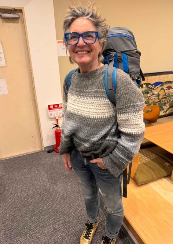 Lächelnde Irmgard Rosina Bauer mit großem Rucksack und blauer Brille – Sinnbild für Aufbruch, Lebensfreude und Aktivität mit 70.