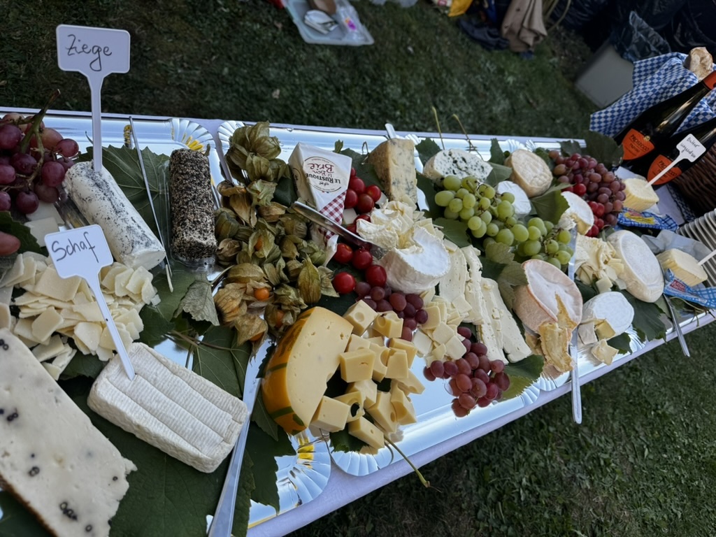 Großes Käsebuffet mit verschiedenen Käsearten, Trauben, Physalis und dekorativen Blättern; kleine Schilder kennzeichnen unter anderem Ziegen- und Schafskäse.