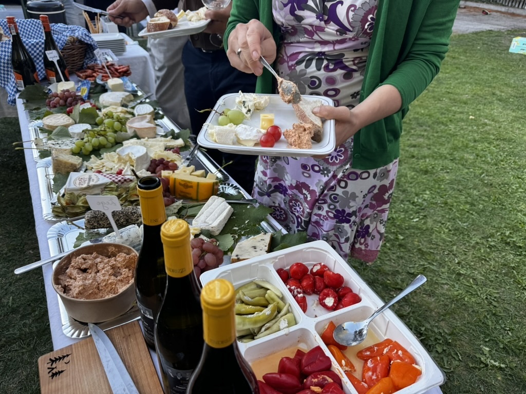 Üppiges Käsebuffet mit verschiedenen Käsesorten, Trauben, eingelegtem Gemüse und Weinflaschen; eine Frau stellt sich gerade einen Teller zusammen.
